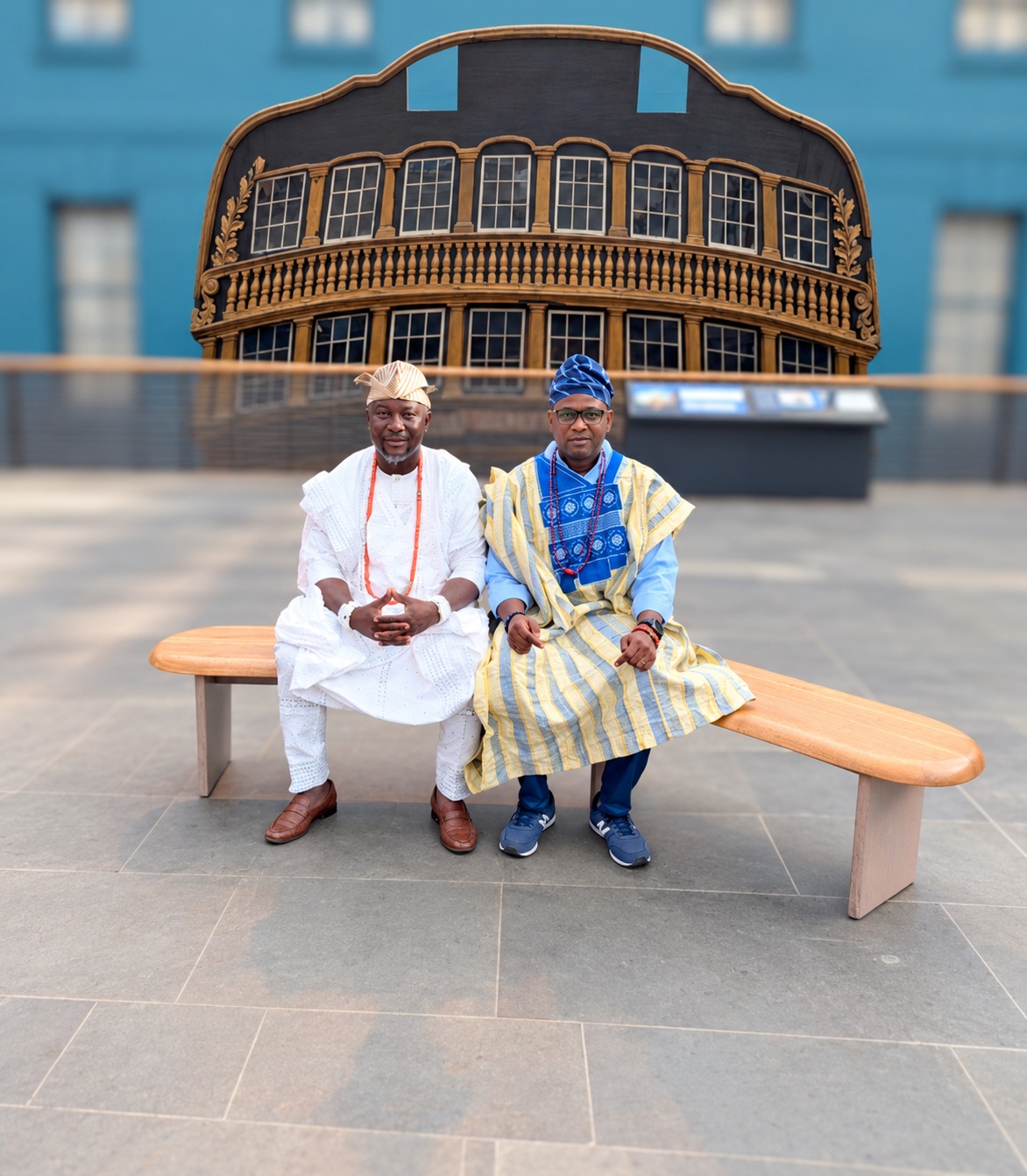 Chief Babatunde Sanni and Ọlálékan Olúkọtún seated in Yorùbá attire before a historic ship display in Greenwich